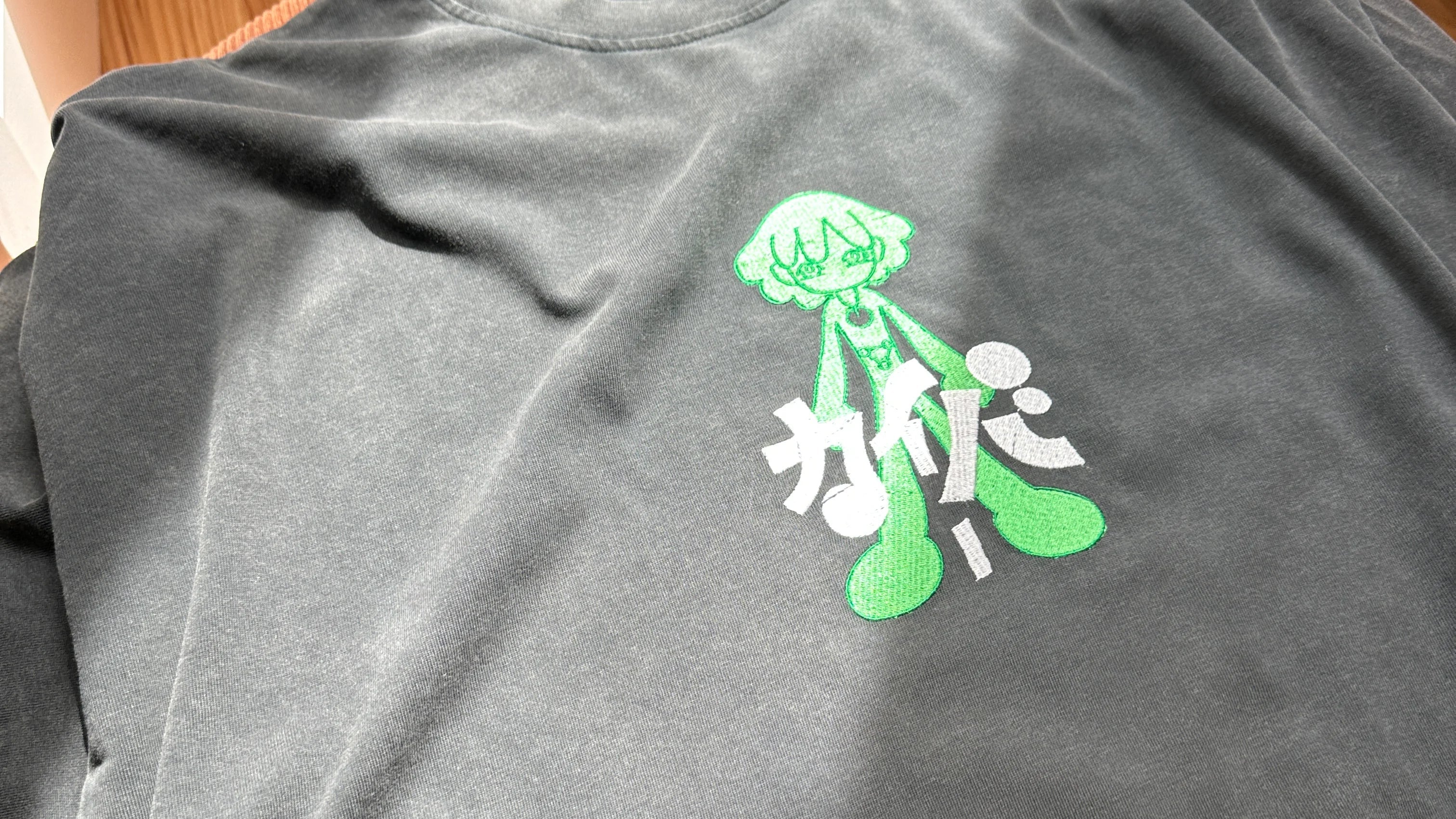 Gray t-shirt with green and white anime embroidery design on a wooden surface